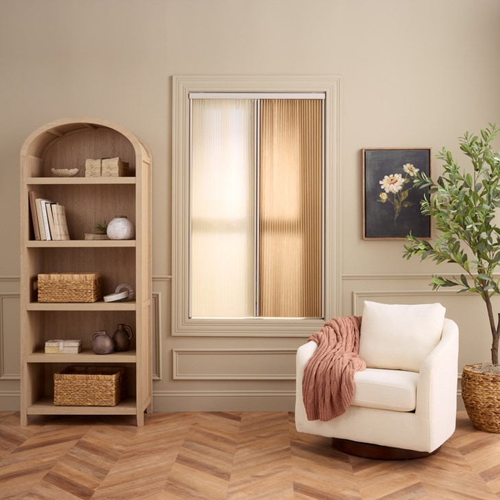 Lifestyle living room shot of a closed Vertical Cellular Shade in the Beach GS2506 color featuring Light Filtering opacity and a Day and Night Split Stack design on a window next to a bookcase and armchair.